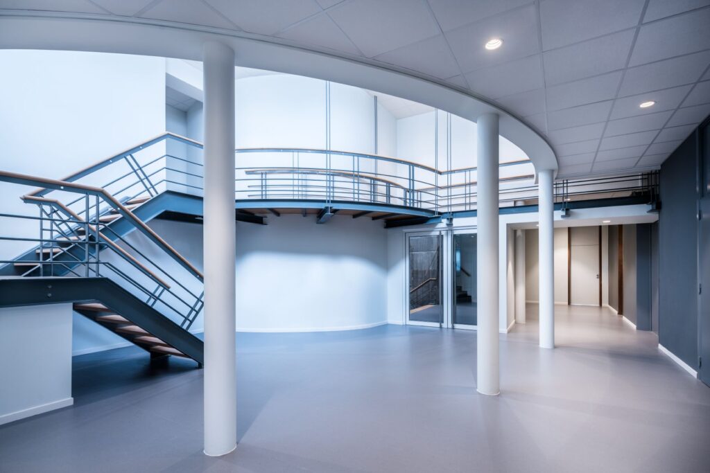 Ljusstark, jämnt upplyst kontorsfoajé med infällda LED-downlights i undertaket som framhäver trappor, pelare och rund balkong och demonstrerar energieffektiva armaturer från PROLJUS AB som skapar en funktionell och modern atmosfär perfekt för kommersiella miljöer