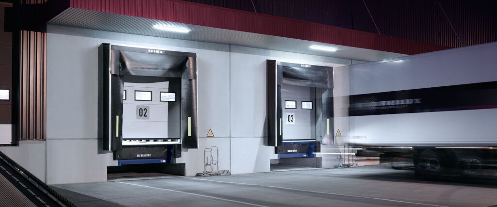 Nattbild av två moderna, energieffektiva PROLJUS LED-armaturer monterade i takets underhäng över lastkajer som ger starkt, jämnt vitt ljus och förbättrar säkerhet och synlighet vid in‑ och utkörning, hållbar och kostnadseffektiv industribelysning från PROLJUS AB