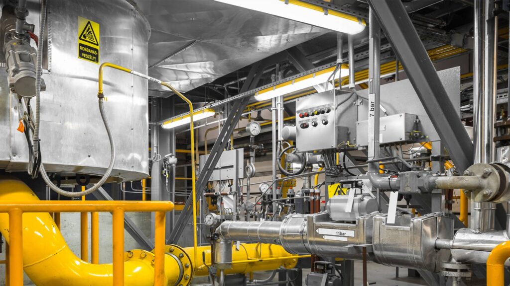 Industriell produktionsmiljö med långa linjära LED-armaturer monterade i tak och över maskiner som ger klart kallvitt, bländfritt ljus och visar PROLJUS AB:s robusta, energieffektiva belysningslösningar för säkra och effektiva arbetsytor