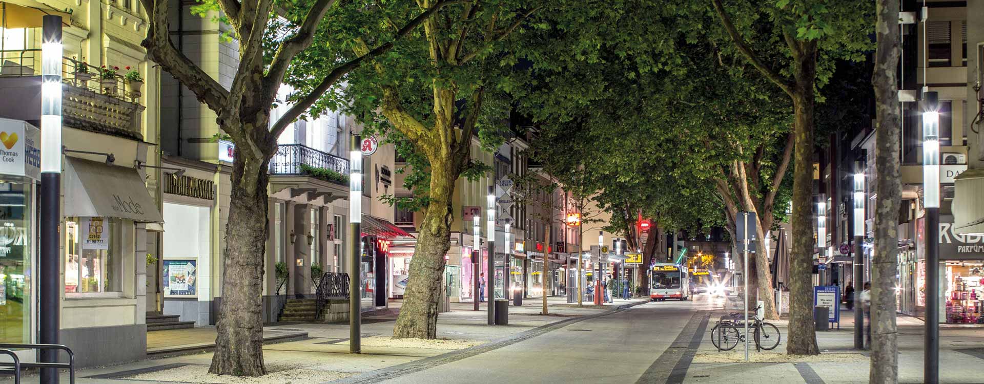 Nattlig gata upplyst av eleganta höga LED-stolpar från PROLJUS AB som ger jämnt, bländfritt ljus mellan träd och butiksfasader och skapar trygga, attraktiva stadsmiljöer perfekta för offentliga och kommersiella projekt