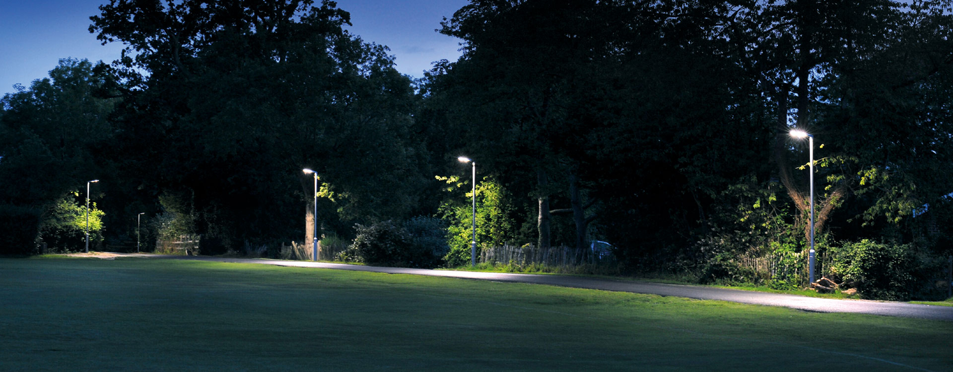 Rad av PROLJUS AB:s eleganta energieffektiva LED‑armaturer längs en parkstig i skymningen, som ger jämnt vitt ljus och framhäver gångväg, träd och grönska — en stilren och robust lösning för kommuner, parker och bostadsområden