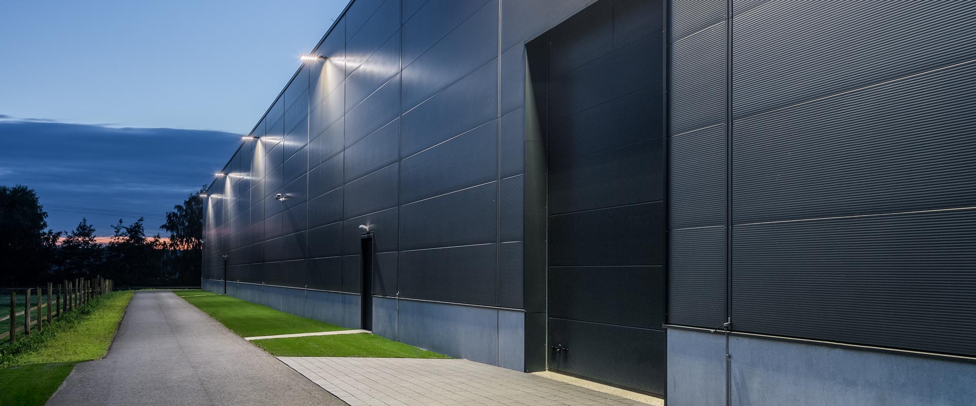 Skymningsbild av en industrifasad upplyst av en rad väggmonterade PROLJUS AB LED-armaturer som ger jämnt, bländfritt och energieffektivt ljus för ökad säkerhet och modern estetik i lager och logistikbyggnader