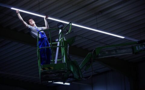 Lång, slank LED-armatur som avger starkt och jämnt vitt ljus i ett industriellt lager, monterad av en tekniker på lift, energieffektiv och idealisk för arbetsplatser, beställ från PROLJUS AB