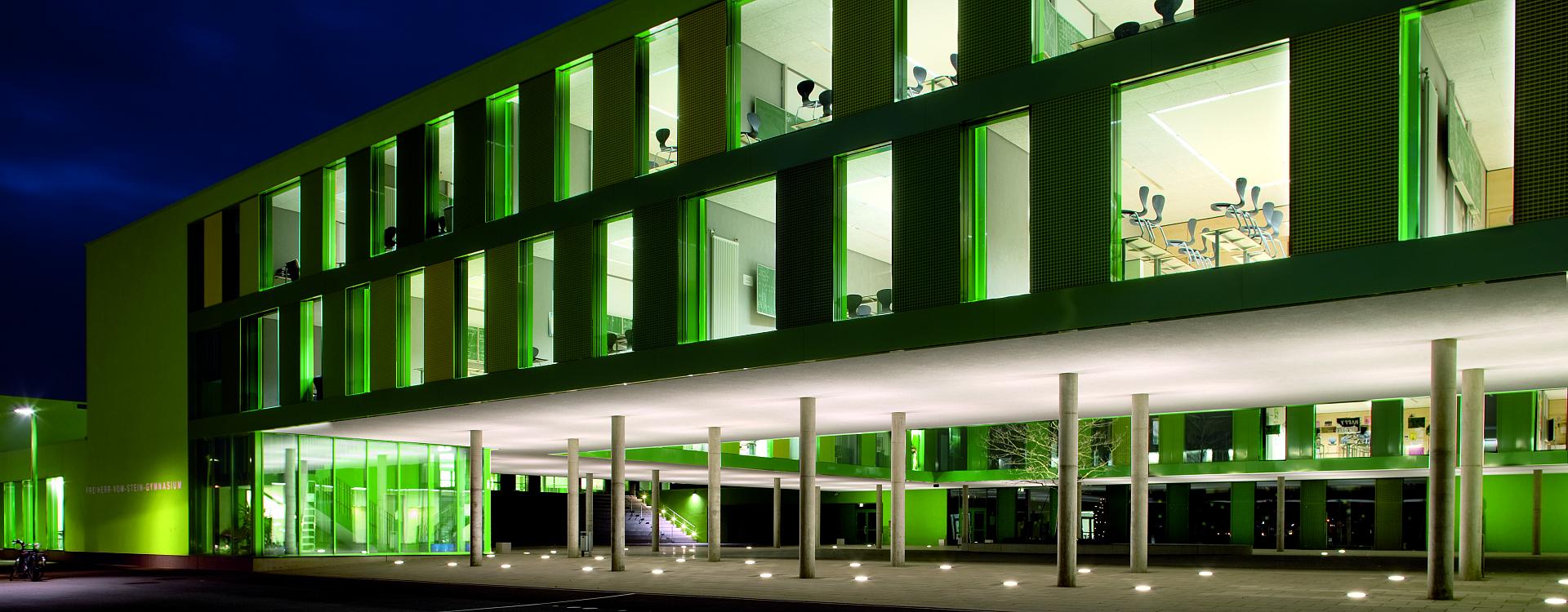 Nattbild av modern byggnad upplyst av PROLJUS ABs LED-armaturer med gröna linjära fasadbelysningar i fönsterspringorna, infällda markspotlights runt entrén och jämna takdownlights under gången som ger energieffektiv, dekorativ och trygg belysning för skolor och offentliga fastigheter