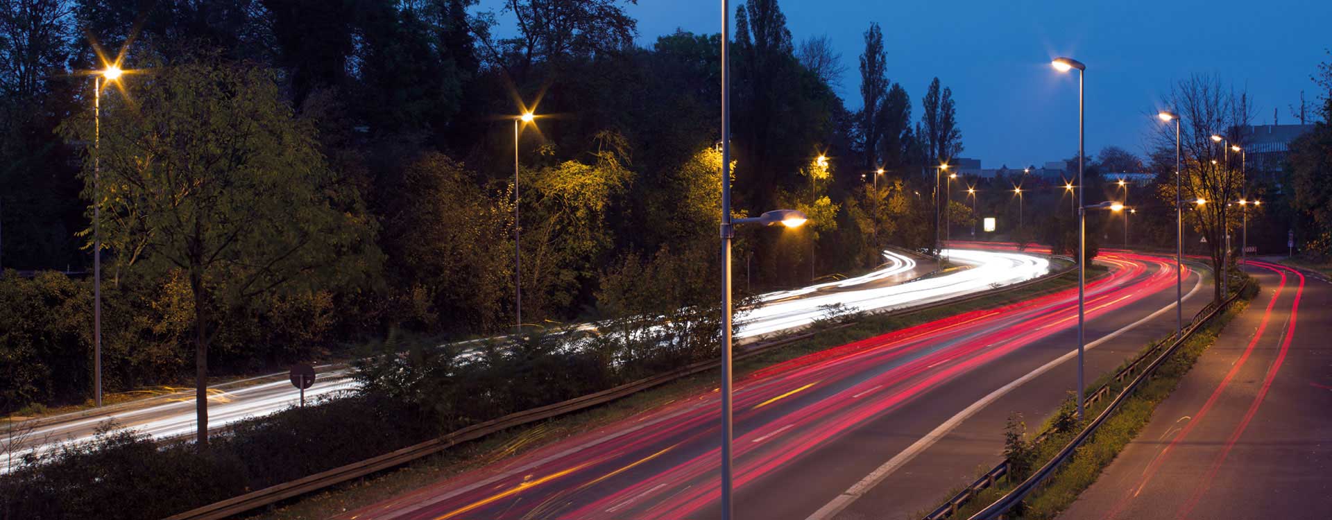 Kvällsbild av motorväg i skymning med en rad moderna, energieffektiva gatlyktor som sprider varmt, jämnt ljus över körfält och framhäver röda och vita ljusspår, en driftsäker och stilren ljuslösning från PROLJUS AB