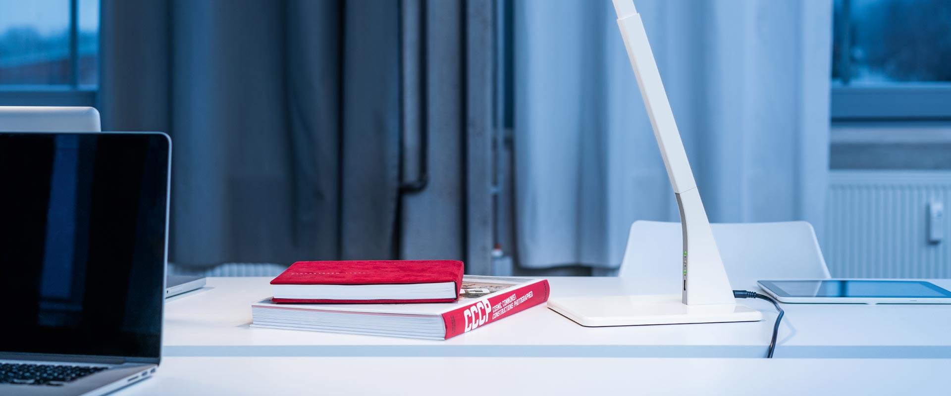 Vit modern LED-skrivbordsarmatur med flexibel arm, inbyggda indikatorer och USB‑port på ett stilrent vitt skrivbord intill laptop och böcker som ger riktat, energieffektivt och ergonomiskt arbetsljus perfekt för kontor och hemmakontor från PROLJUS AB