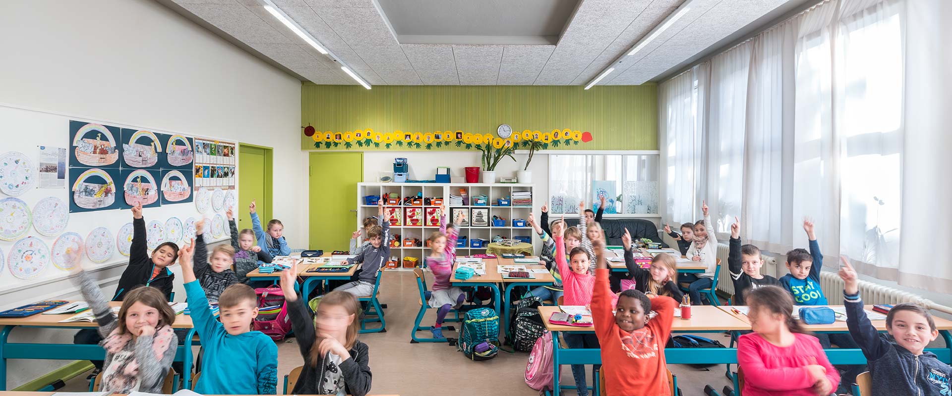 Klassrum med barn som räcker upp händerna, jämnt upplyst av moderna linjära infällda LED-armaturer i taket och dagsljus genom stora fönster, demonstrerar PROLJUS ABs energieffektiva bländfria skolbelysning som förbättrar arbetsmiljön och sparar energi