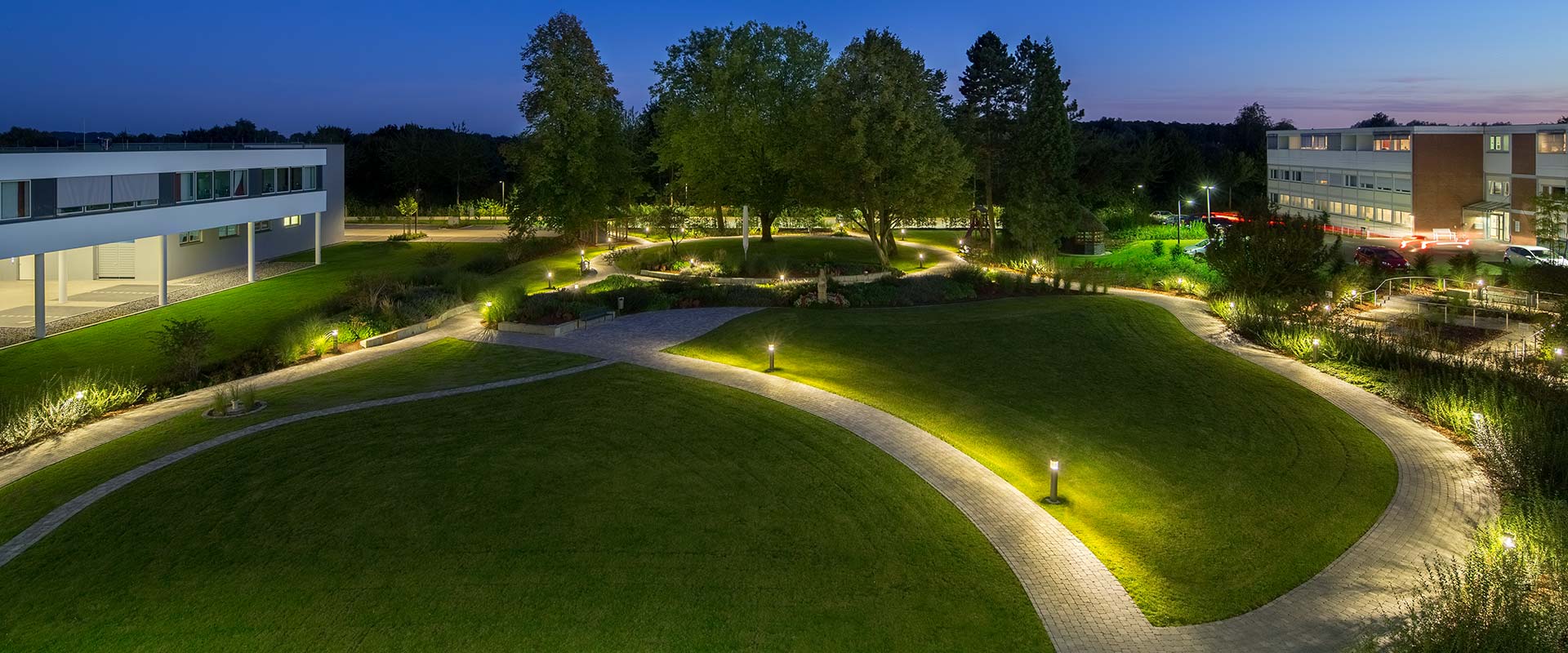 Kvällsbild av en park med slingrande gångvägar kantade av moderna pollare och markbelysning från PROLJUS AB som ger jämnt, varmt ljus och trygg vägvisning vid gräskullar, planteringar och byggnadsentréer