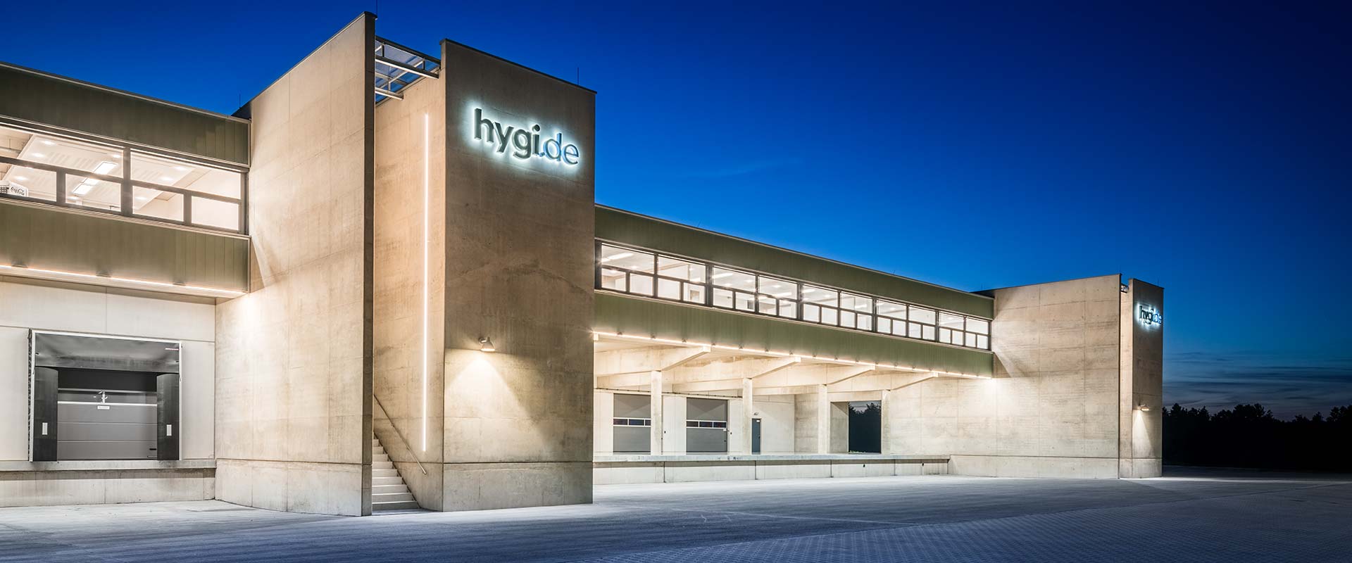 Nattbild av modern lager- och logistikbyggnad där PROLJUS AB:s energieffektiva LED-armaturer och linjära ljusräcken markerar rampen och fasaden med jämnt vitt ljus, framhäver betongens struktur, ökar säkerheten vid lastning och erbjuder stilren kostnadseffektiv belysning som är idealisk för industriella anläggningar