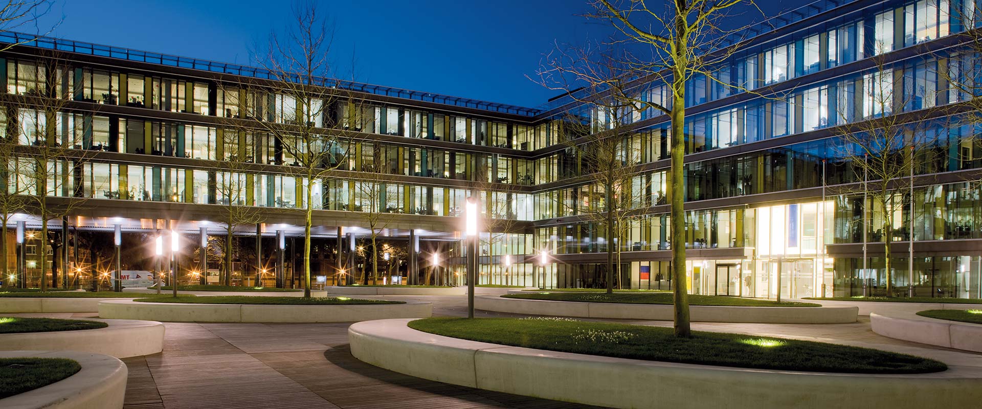 Nattbild av modern kontorsinnergård upplyst av eleganta PROLJUS AB-stolplampor som ger varm, jämn och energieffektiv belysning, framhäver glasfasaden, planteringar och gångstråk och skapar en inbjudande företagsmiljö