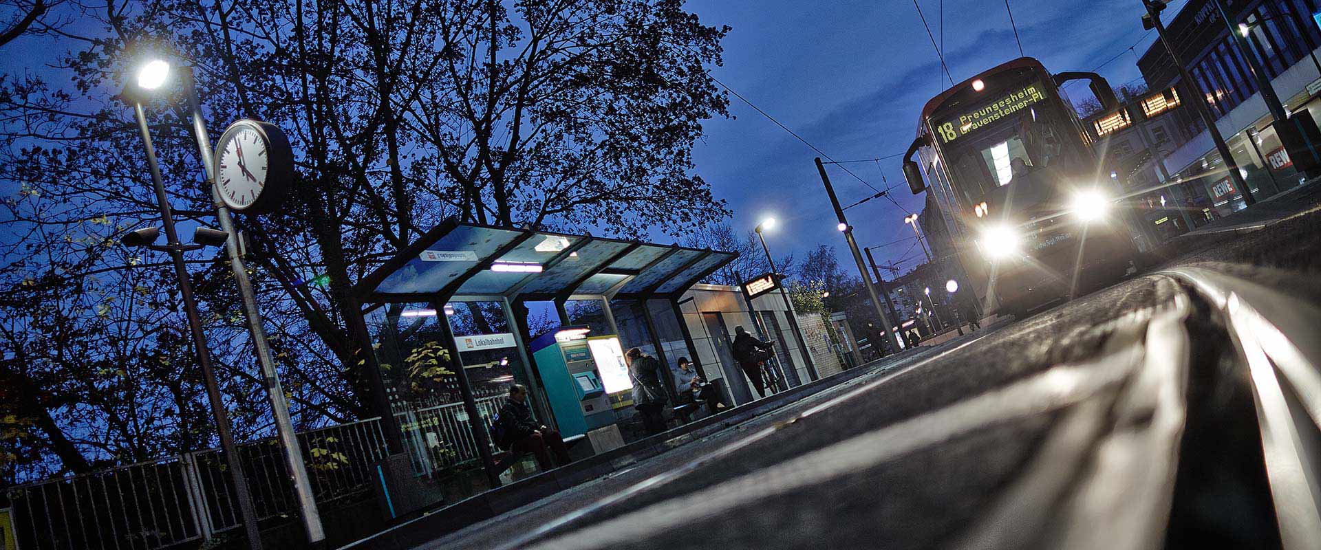 Kvällsbild av en spårvagnshållplats med kraftfulla gatlyktor takmonterade LED armaturer i väntkuren och starka strålkastare från vagnen som lyser upp plattformen Visar PROLJUS ABs energieffektiva robusta och driftssäkra belysningslösningar som ökar trygghet och förbättrar stadsmiljön