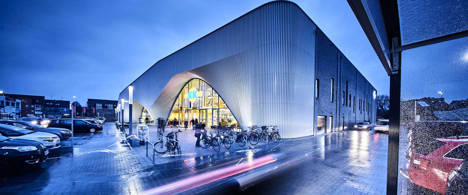 Kvällsbild av en modern butik med våt entré och reflekterande mark, elegant upplyst av PROLJUS AB:s energieffektiva utomhusarmaturer som framhäver byggnadens arkitektoniska valv, lyser upp cykelparkeringen och parkeringsplatsen med jämnt vitt ljus och skapar en inbjudande atmosfär som ökar kundflödet