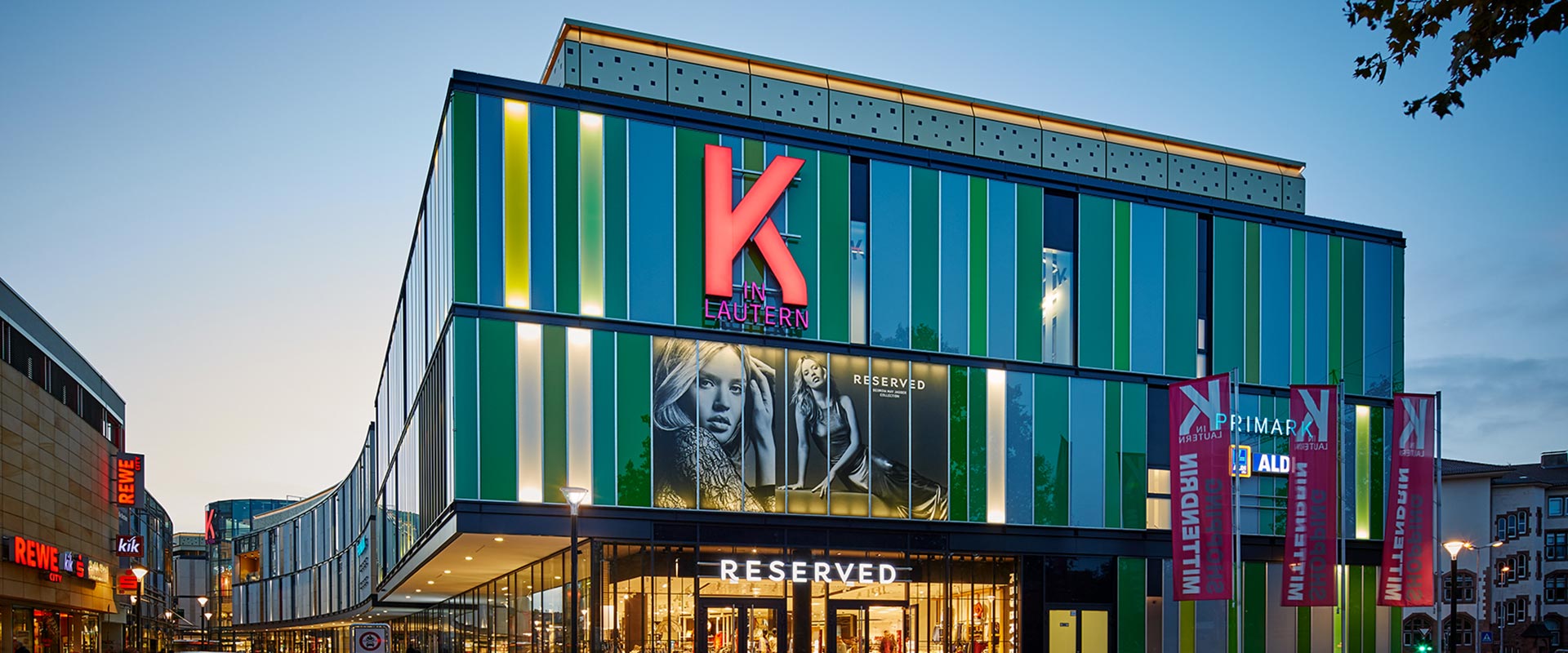 Skymningsvy över ett köpcentrum där energieffektiva fasadarmaturer och gatlyktor från PROLJUS AB framhäver färgglada fasadpaneler, stora skyltar och butiksentréer och skapar en inbjudande kvällsatmosfär som ökar kundflödet