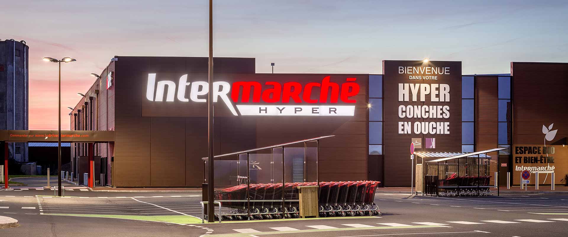 Kundparkering vid en Intermarché i skymning belyst av höga, moderna LED-armaturer från PROLJUS AB som sprider varmt, jämnt ljus över fasad, kundvagnar och gångbanor och framhäver butiksprofilen samtidigt som energieffektiv, robust design och låg driftkostnad gör dem till en kostnadseffektiv lösning för detaljhandeln