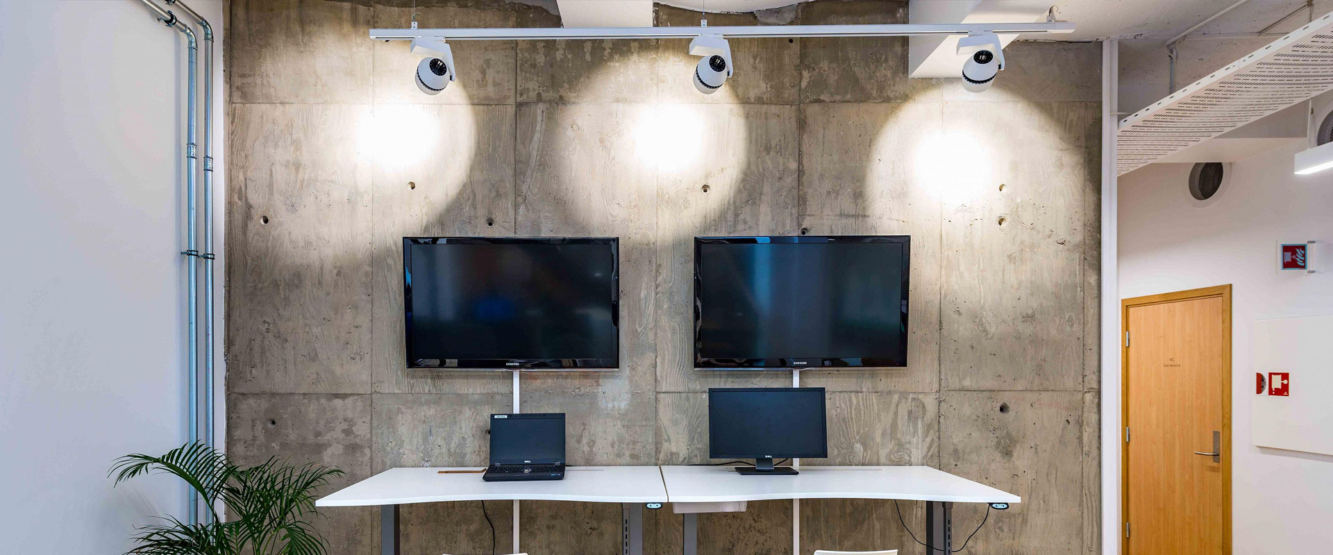 Tre vita spothuvuden monterade på en skena kastar jämna, fokuserade ljusfläckar på en rå betongvägg ovanför två skärmar och visar PROLJUS ABs energieffektiva och stilrena armaturer för kontor och utställningar