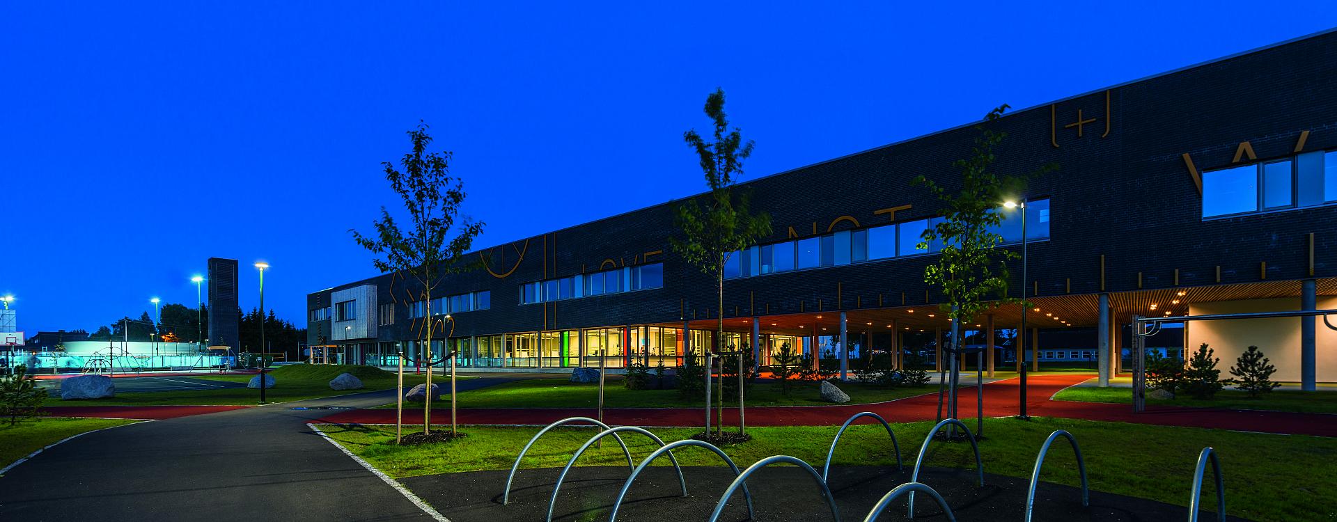 Nattbild av modern byggnad och idrottsplats upplysta av PROLJUS ABs armaturer, höga LED-sportbelysningar ger kraftigt bländfritt ljus över planen, pollare och infällda takarmaturer skapar varm och trygg atmosfär längs gångvägar och fasad, energieffektiva hållbara lösningar som lyfter säkerhet och estetik för skolor kommuner och fastighetsägare