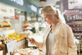 Kund i matbutik tittar på mobil under moderna, energieffektiva PROLJUS AB LED-armaturer som ger klart, färgkorrigerat ljus över frukt och hyllor och skapar en inbjudande butiksmiljö som ökar försäljningen