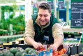 En glad butiksperson placerar tomater under klart vitt ljus från PROLJUS ABs effektiva butiksarmaturer som framhäver färgerna och skapar en inbjudande miljö för ökad försäljning