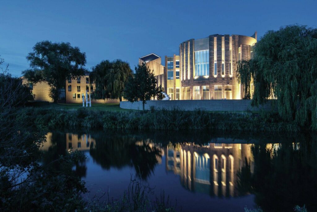 Nattfoto av en samtida byggnad vid sjö med varm fasadbelysning som speglas i vattnet, elegant och energieffektiv utomhusbelysning levererad av PROLJUS AB som framhäver arkitektur och omgivande träd