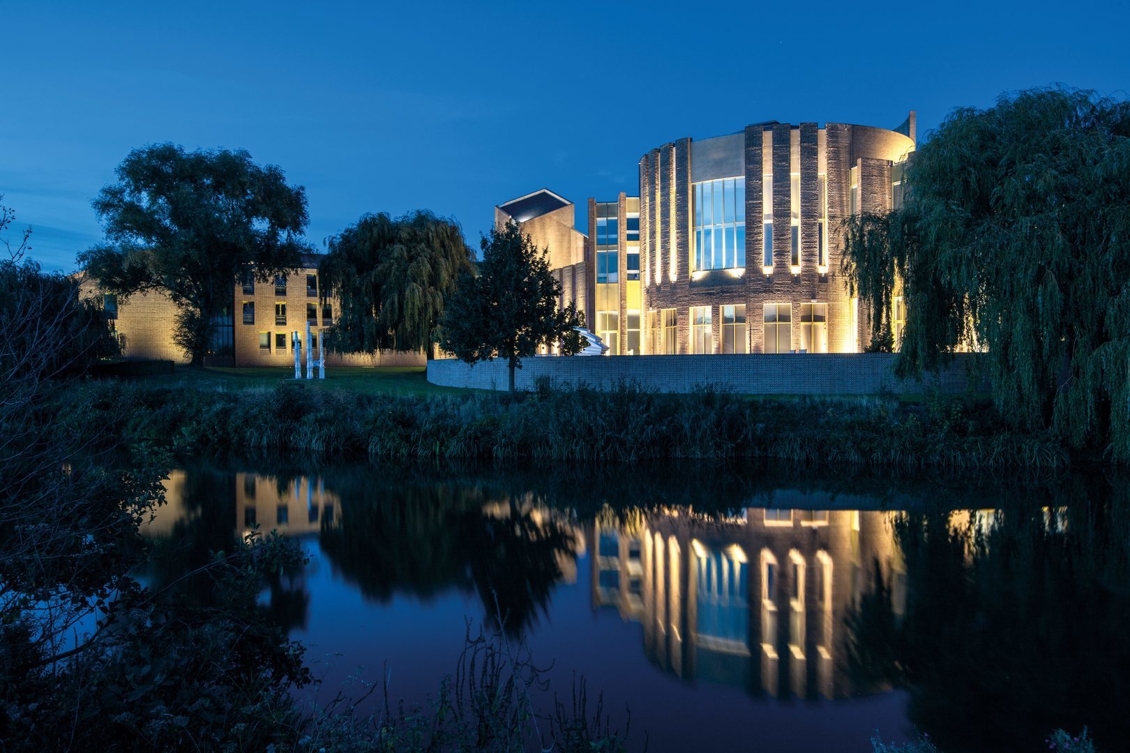 Kvällsbild av en modern byggnad där varma, riktbara och energieffektiva utomhusarmaturer från PROLJUS AB lyfter fram fasadens pelare och stora glasytor, speglas i vattnet och visar hur professionell fasadbelysning ökar estetik och värde