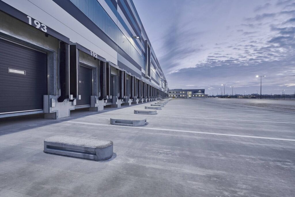 Stort modernt logistikområde i skymning upplyst av jämnt fördelade stolparmaturer som ger klart vitt, energieffektivt LED-ljus över lastkajer och parkeringar, robust industriell konstruktion och lång räckvidd som visar pålitliga belysningslösningar från PROLJUS AB