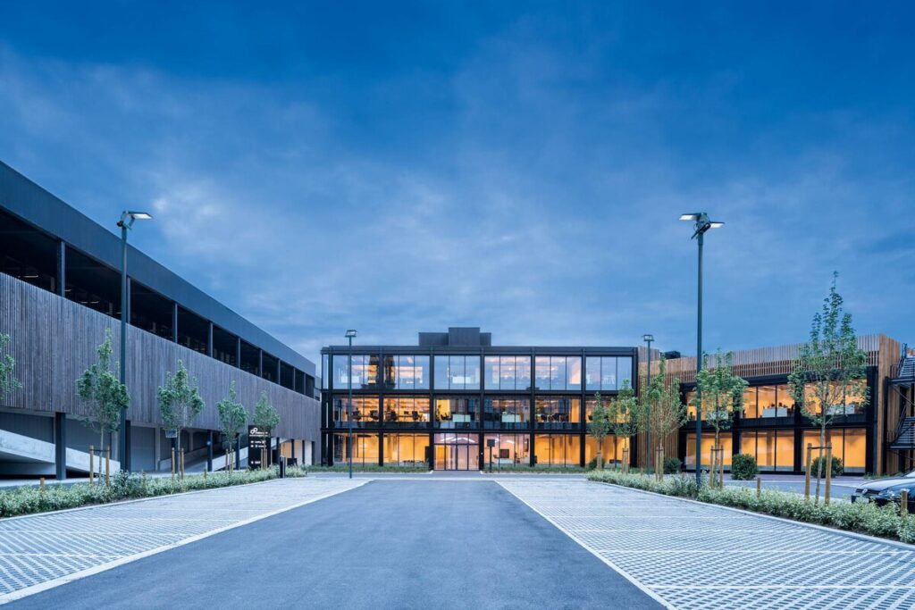 Skymningsbild av modern kontorsbyggnad och parkeringsyta upplyst av PROLJUS AB:s energieffektiva pollararmaturer och varmvita fasadarmaturer, som markerar infart, skapar trygghet och framhäver glasfasaden, perfekt för företag som vill höja säkerhet och profil