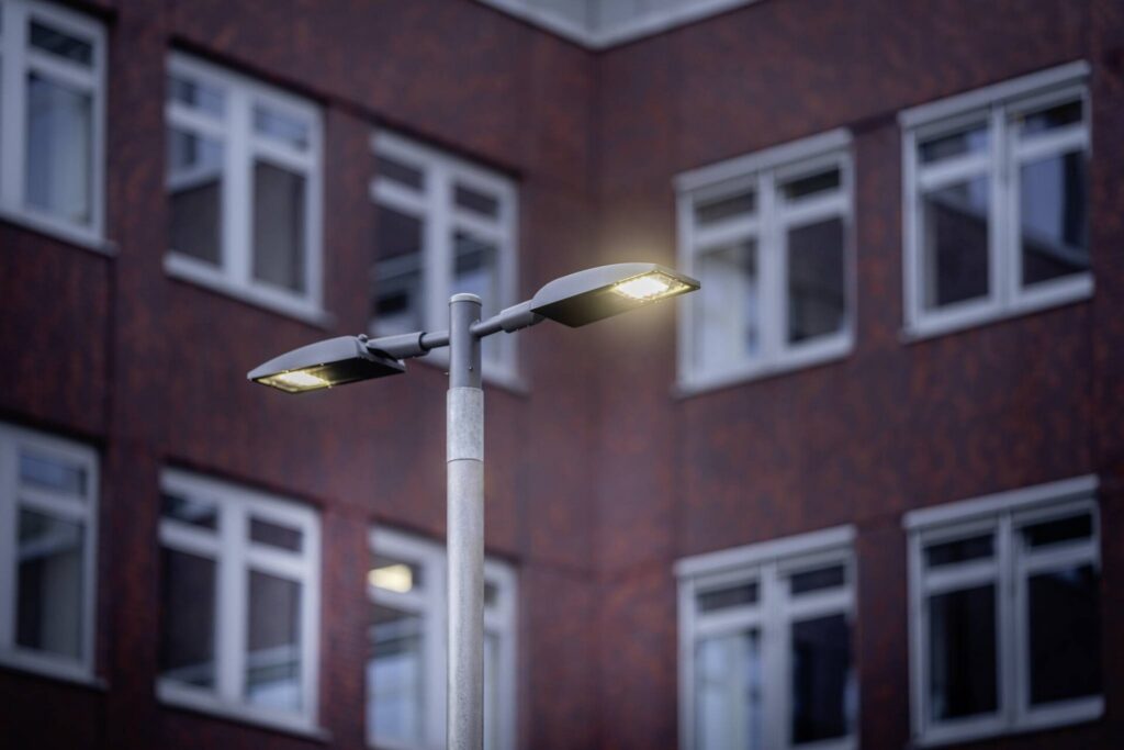 Tvåarmad modern LED-gatubelysning på metallstolpe som avger varmt, energieffektivt sken framför kontorsfasad, robust och stilren armatur från PROLJUS AB idealisk för urbana miljöer