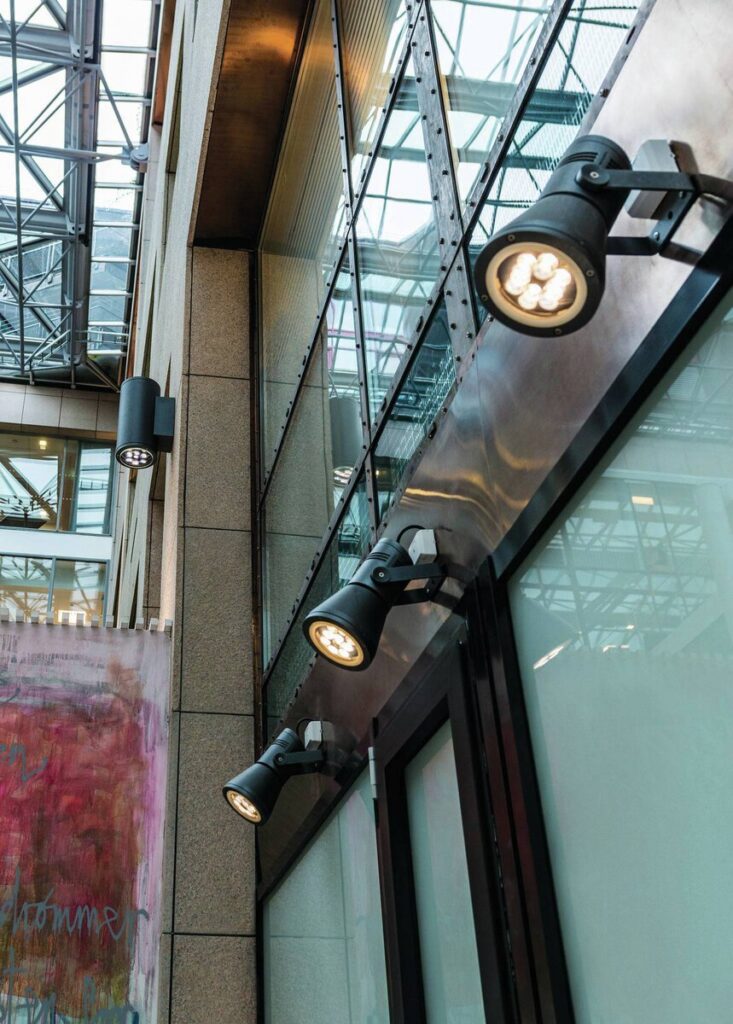 Flera svarta, riktbara LED-strålkastare från PROLJUS AB monterade på glas- och metallfasad i ett atrium, varmvit riktad belysning som framhäver arkitektur och skyltfönster, energieffektiva och robusta lösningar som ökar synlighet och attraktivitet i kommersiella miljöer