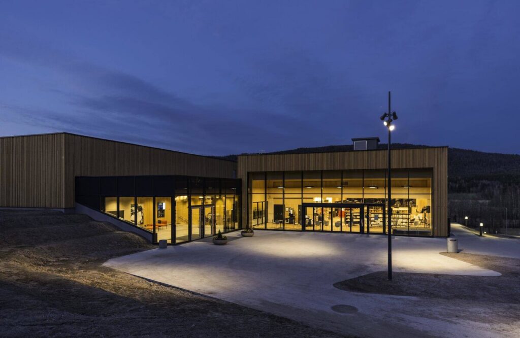 Skymningsbild av modern träbyggnad där PROLJUS ABs eleganta utomhusarmaturer och stolpbelysning lyser upp entré och fasad med varmt energieffektivt ljus och visar en hållbar säljande lösning för offentliga miljöer