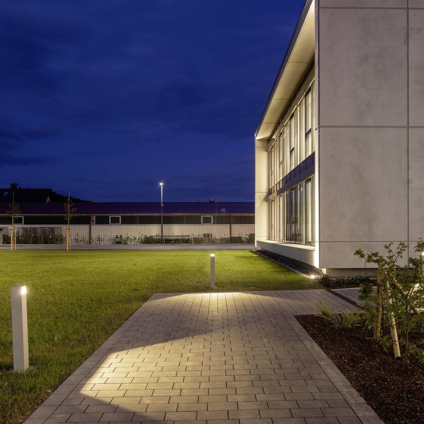Kvällsbild av modern byggnadsfasad och gångväg upplyst av diskreta, energieffektiva pollare och varm LED-fasadbelysning från PROLJUS AB som skapar jämn, säker och estetisk utomhusbelysning idealisk för kommersiella och bostadsprojekt