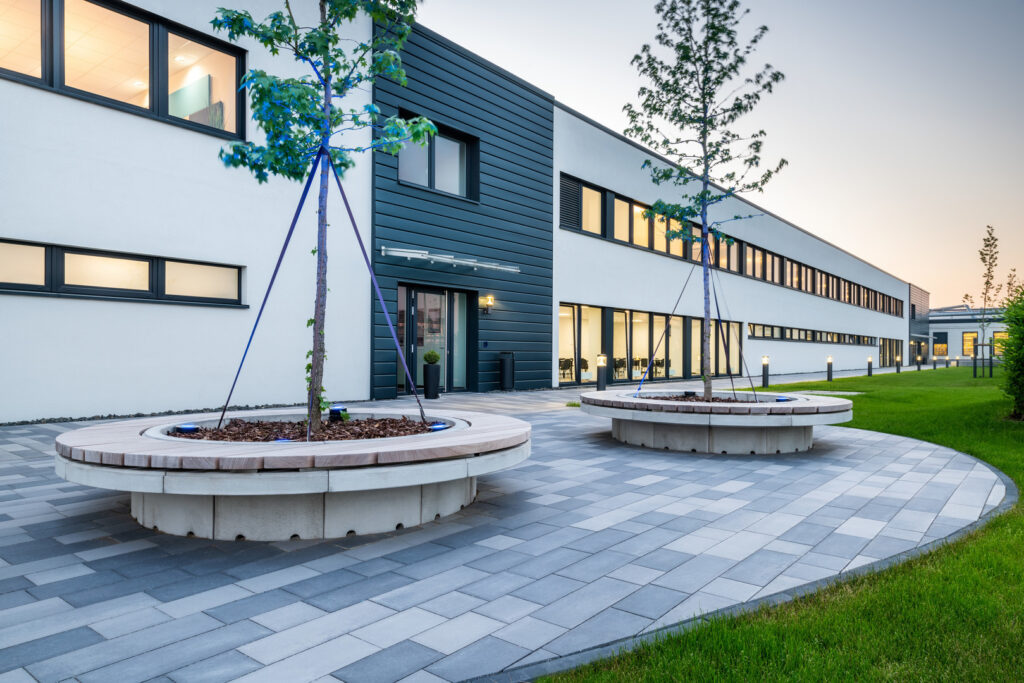 Modern kontorsfasad kvällstid belyst av blå uplights runt cirkulära planteringar och varmvita pollare längs gångstråket perfekt utomhusbelysning från PROLJUS AB
