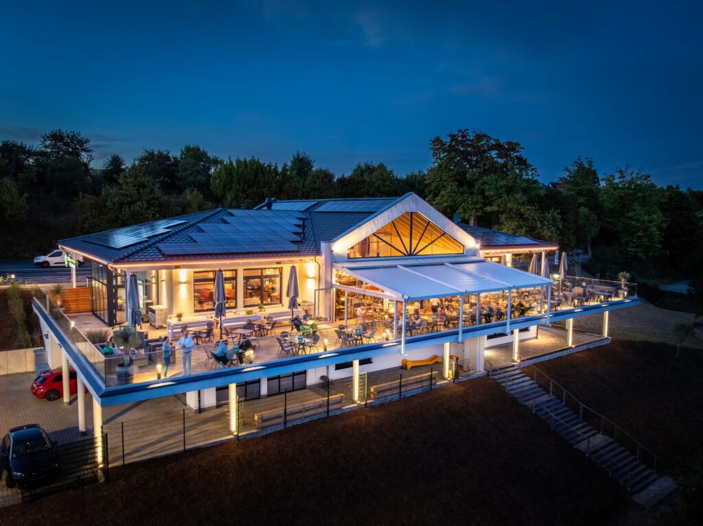 Modern restaurang med belyst uteterrass i skymning, panoramavy över uteservering och glasad pergola på höjd.