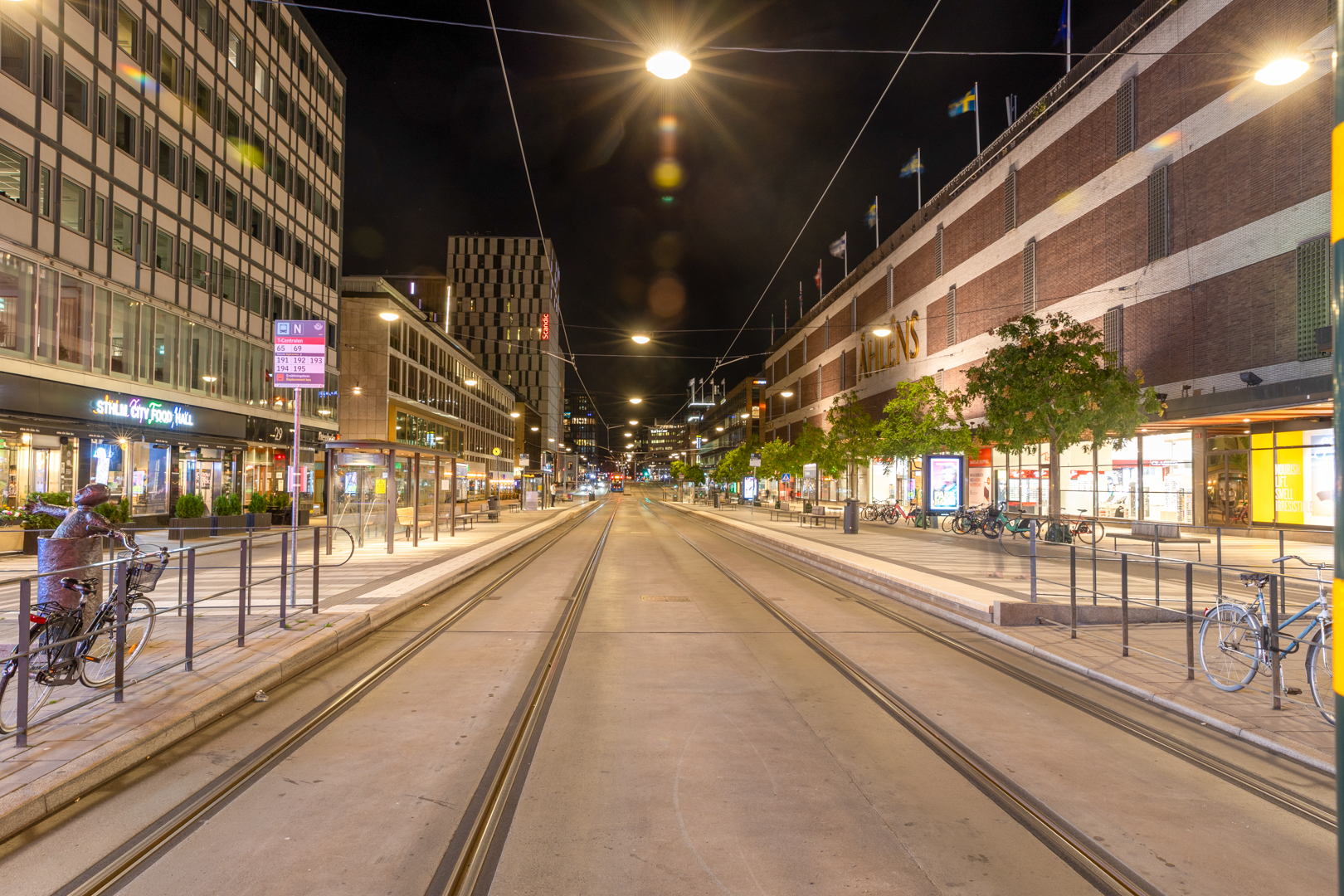Centrala Stockholm nattetid med spårvagnsspår, Åhléns varuhus, upplysta butiker, cyklar och hållplatser.