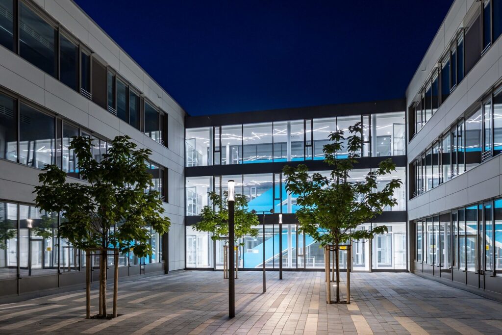 Nattbild av en modern kontorsinnergård upplyst av slanka pollare och linjära innertaksarmaturer som skapar jämn och energieffektiv belysning samt framhäver byggnadens arkitektur, en designad belysningslösning från PROLJUS AB