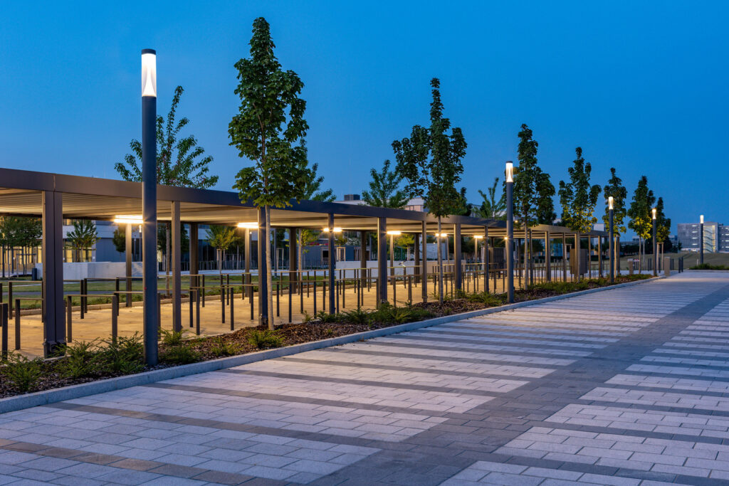 Kvällsbild av en offentlig gånggata och pergola upplyst av PROLJUS ABs slanka LED-armaturer på stolpar som ger varmt, jämnt sken och framhäver arkitektur och stenläggning, idealisk för trygg och energieffektiv stadsmiljö