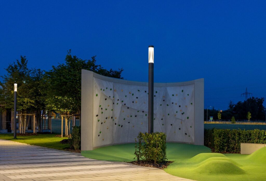 Två stilrena svarta pollarljus från PROLJUS AB avger mjukt varmvitt LED-ljus som elegant lyser upp en kvällspark, framhäver en klättervägg och gångväg och visar energieffektiv design och hög kvalitet idealisk för offentliga miljöer