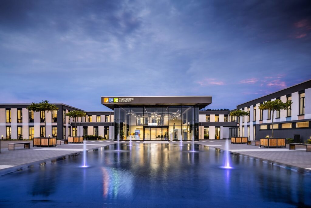 Kvällsbild av modernt kontorskomplex belyst med PROLJUS ABs energieffektiva LED-armaturer, varmvitt fokusljus i glasentrén, fasad- och trädspotlights samt violetta fontänstrålar och markbelysning som speglas i vattenspeglingen och skapar en inbjudande atmosfär