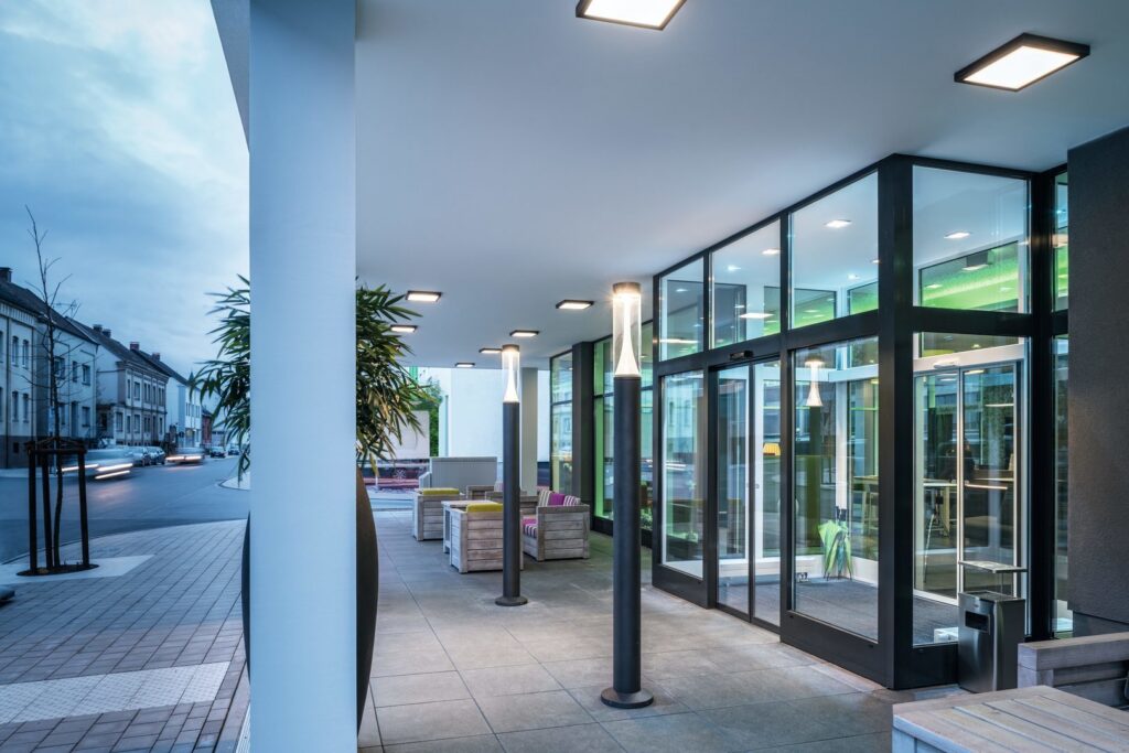 Modern entrébelysning från PROLJUS AB med slanka pollarlampor och infällda takarmaturer som avger jämnt, varmvitt LED-ljus och skapar en inbjudande, energieffektiv och stilren atmosfär vid glasentrén perfekt för hotell och kontor