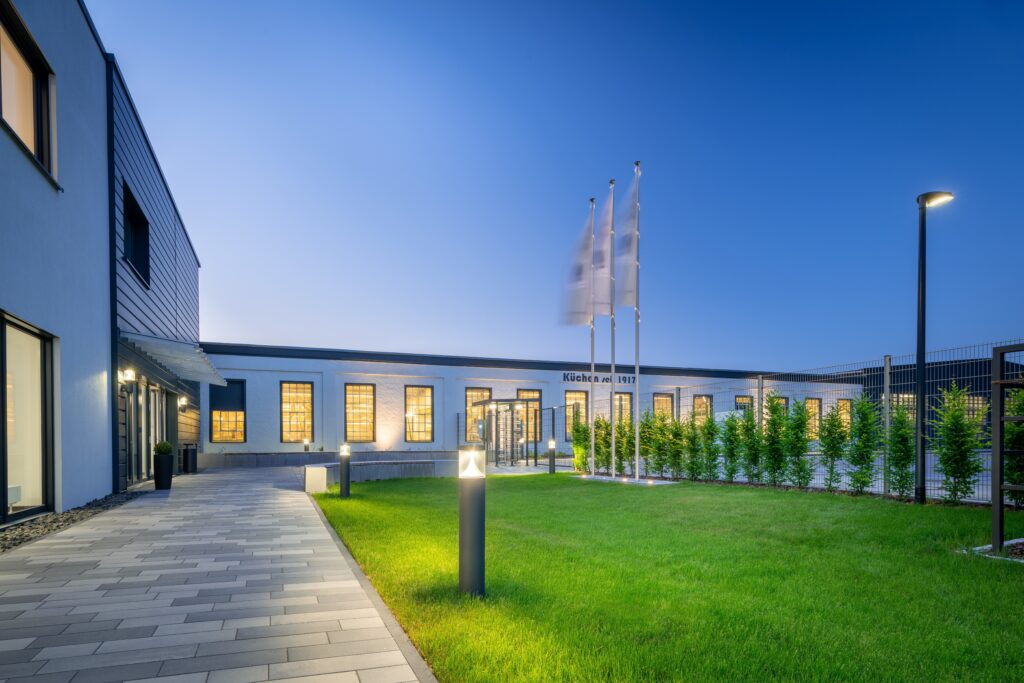 Kvällsbild där PROLJUS ABs eleganta pollare och stolpar i mörk finish sprider varmt, jämnt LED-ljus över gångväg och gräsmatta, framhäver fasaden, ökar tryggheten och visar på energieffektiv, hållbar utomhusbelysning perfekt för företag