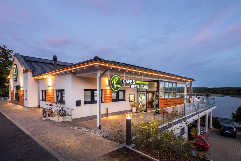 Modern café och restaurang vid sjö, kvällsbelysning och uteservering på terrass med utsikt över vattnet.