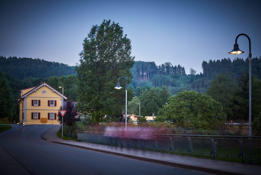 Kvällsbild med två svanhalsarmaturer från PROLJUS AB som avger varmvit LED-belysning över en bro och trottoar, med robusta stålstolpar och stilren design perfekt för småstadsgator