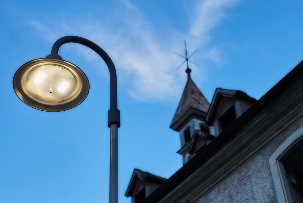 Närbild av en upplyst gatubelysningsarmatur från PROLJUS AB med rund reflektor och varm, energieffektiv ljuskälla mot blå kvällshimmel och äldre byggnad i bakgrunden, en elegant och hållbar lösning för stads- och miljöanpassad belysning