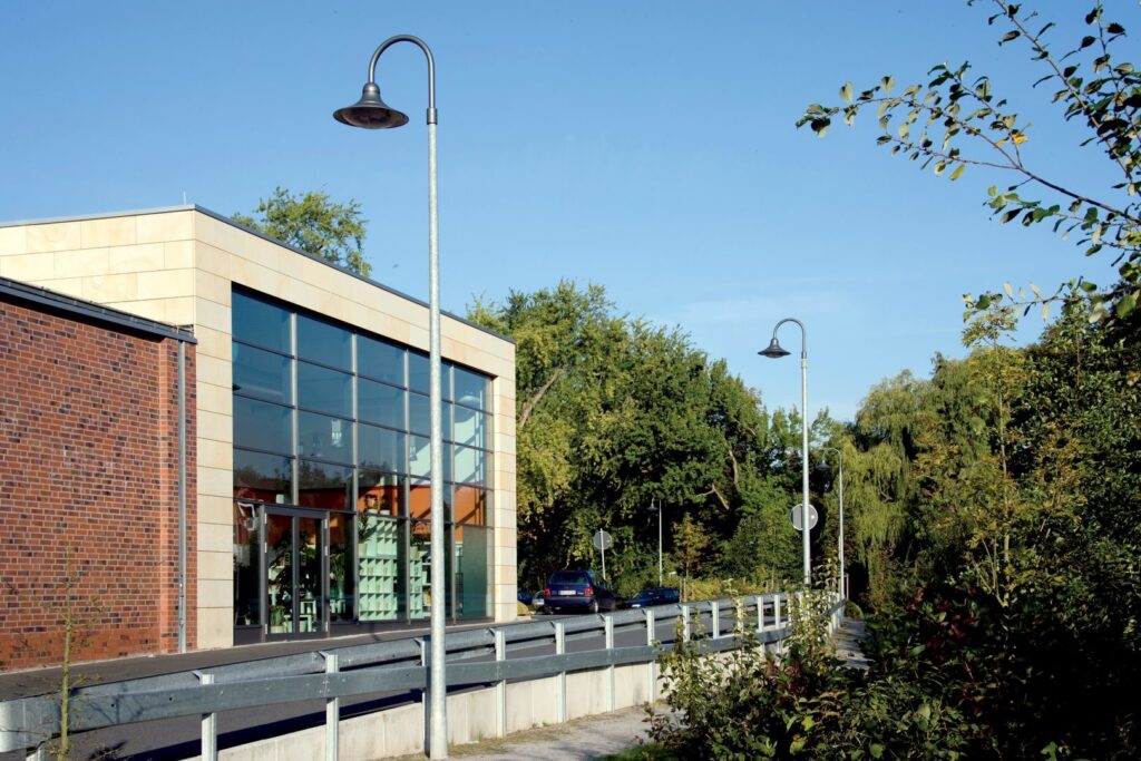 Elegant svängd gatlykta från PROLJUS AB längs trottoar och väg framför en glasbyggnad med varm nedåtriktad ljusbild, robust metallstolpe och tidlös design som förhöjer trygghet och estetik i offentliga miljöer