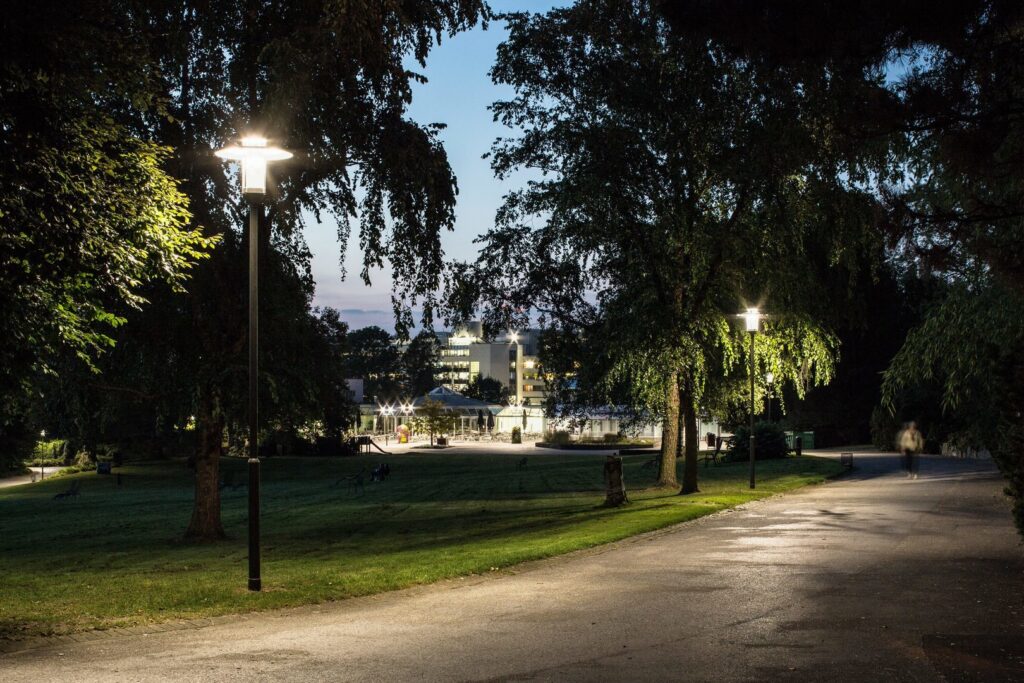 Kvällsbild i park med PROLJUS ABs moderna, energieffektiva LED-armaturer på smala svarta stolpar som ger bländfritt, varmt ljus över gångväg och gräsmatta och framhäver trädens lövverk perfekt för trygg och attraktiv utomhusbelysning