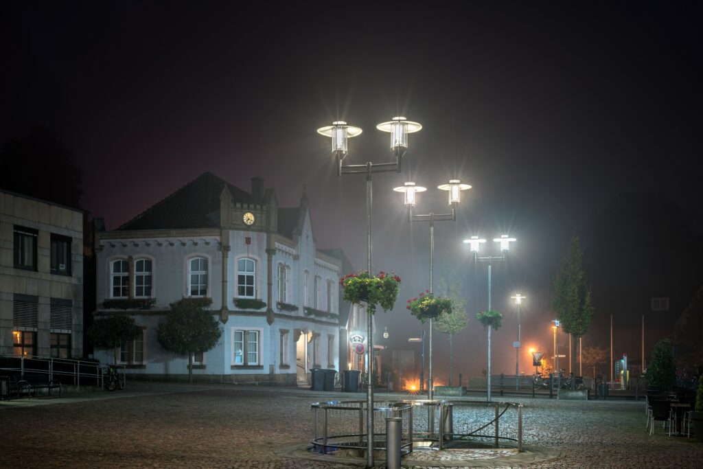 Nattligt torg belyst av flera moderna gatlyktor med skivformade skärmar och bländfritt LED-ljus samt hängande blomkorgar, en energieffektiv och dekorativ lösning för offentliga rum från PROLJUS AB