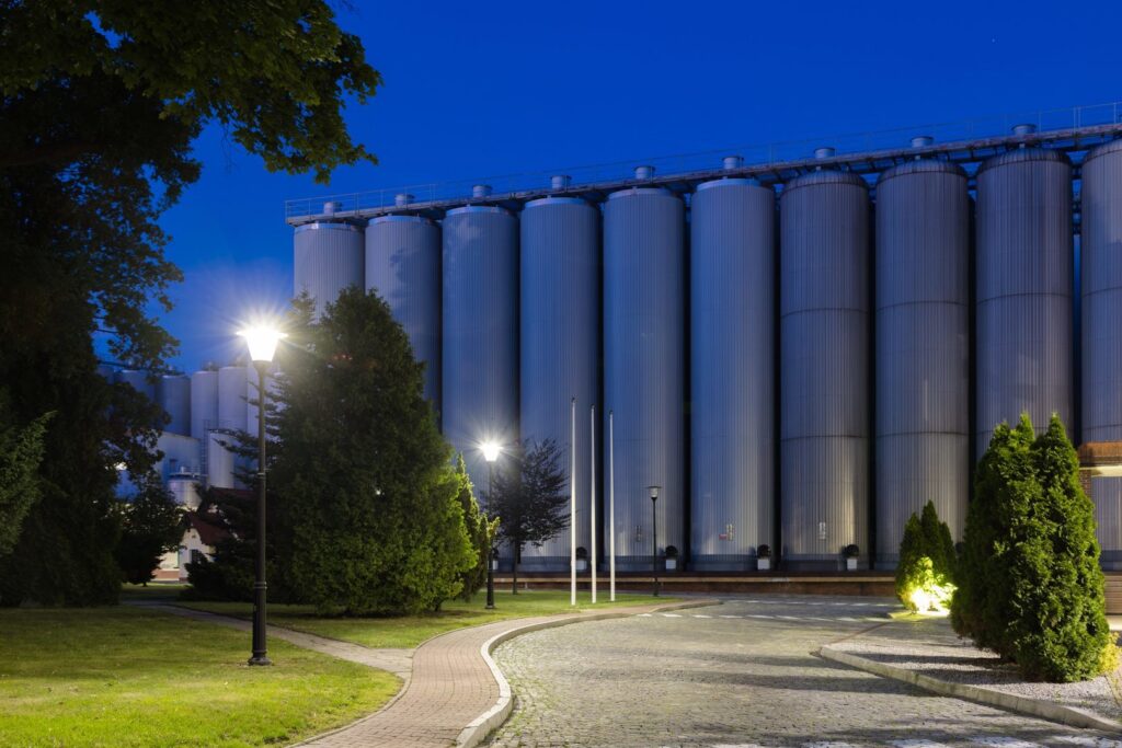 Kvällsbild av industrimiljö där PROLJUS AB-gatlampor lyser upp en gångväg, gräsytor och höga silos med klart, jämnt ljus, energieffektiv, modern design som framhäver säkerhet och estetik i fabriks- och parkområden