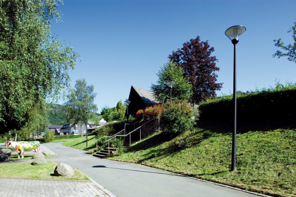 Solbelyst parkväg med flera slanka svarta gatlyktor och parkarmaturer från PROLJUS AB med klara kupor som ger jämn, energieffektiv belysning av gångväg och trappa, en stilren lösning för offentliga miljöer