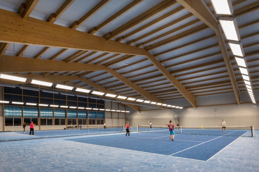 Inomhus tennishall med robusta avlånga LED-armaturer från PROLJUS AB monterade längs träbalkar som ger jämnt, bländfritt och energieffektivt ljus över banorna och förbättrar spelkomfort och driftsbesparingar