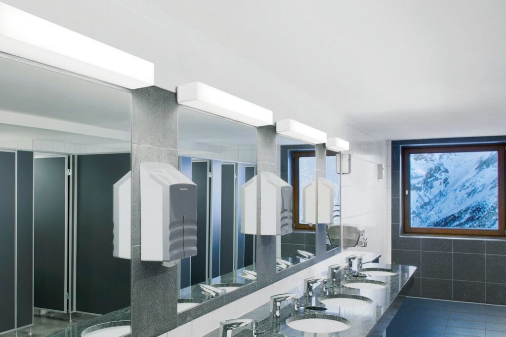 Väggmonterade rektangulära LED-armaturer från PROLJUS AB ovanför speglar i ett modernt offentligt badrum, ger jämn, bländfri och energieffektiv belysning i stilren design som förbättrar komfort och estetik i kommersiella miljöer