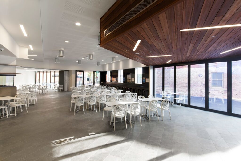 Rymlig cafeteria upplyst av PROLJUS ABs ljuslösningar med infällda cirkulära downlights i vit takyta och slanka linjära LED-lister i träklädd nisch som ger jämn, energieffektiv och modern belysning perfekt för kommersiella miljöer